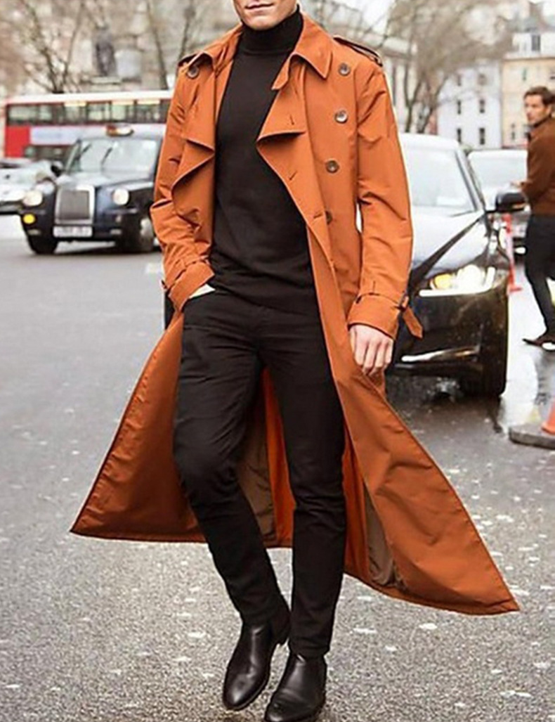 Uomo con trench coat marrone, maglione nero e pantaloni neri, cammina in strada urbana. Moda autunnale, stile elegante, abbigliamento maschile.
