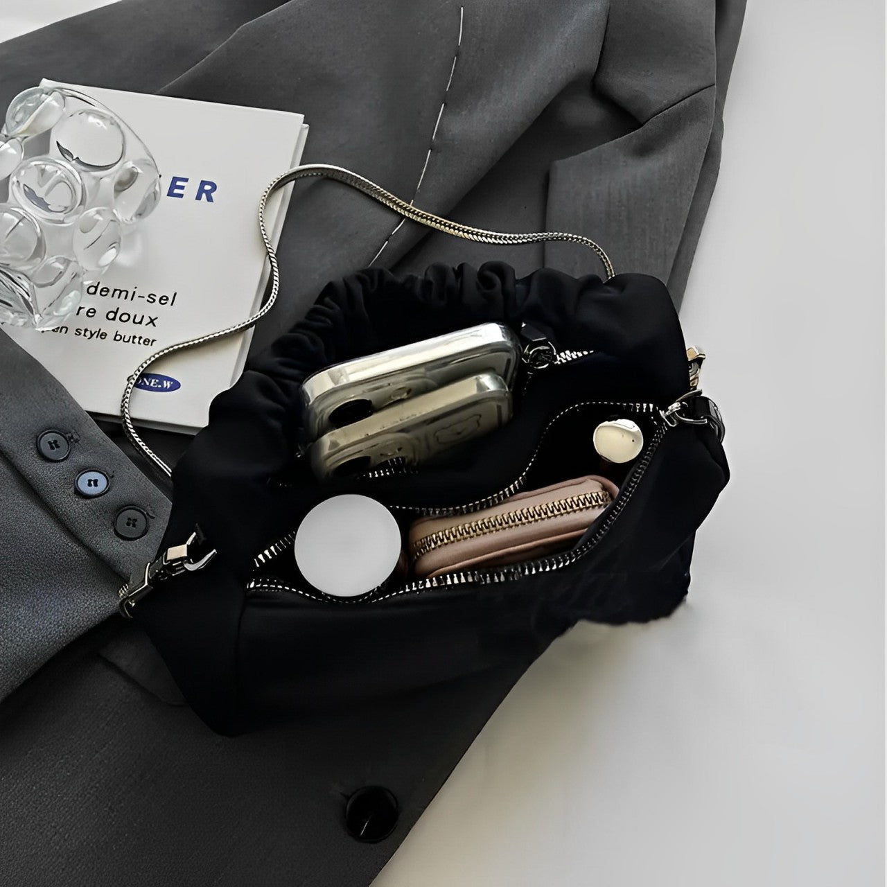 Borsa a tracolla nera in raso con catena argento, contenente smartphone, portafoglio e accessori, su giacca grigia. Elegante e pratica.