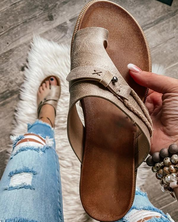 Sandali in pelle marrone chiaro con dettagli intrecciati, tenuti in mano su sfondo di pavimento in legno e tappeto bianco, jeans strappati visibili.
