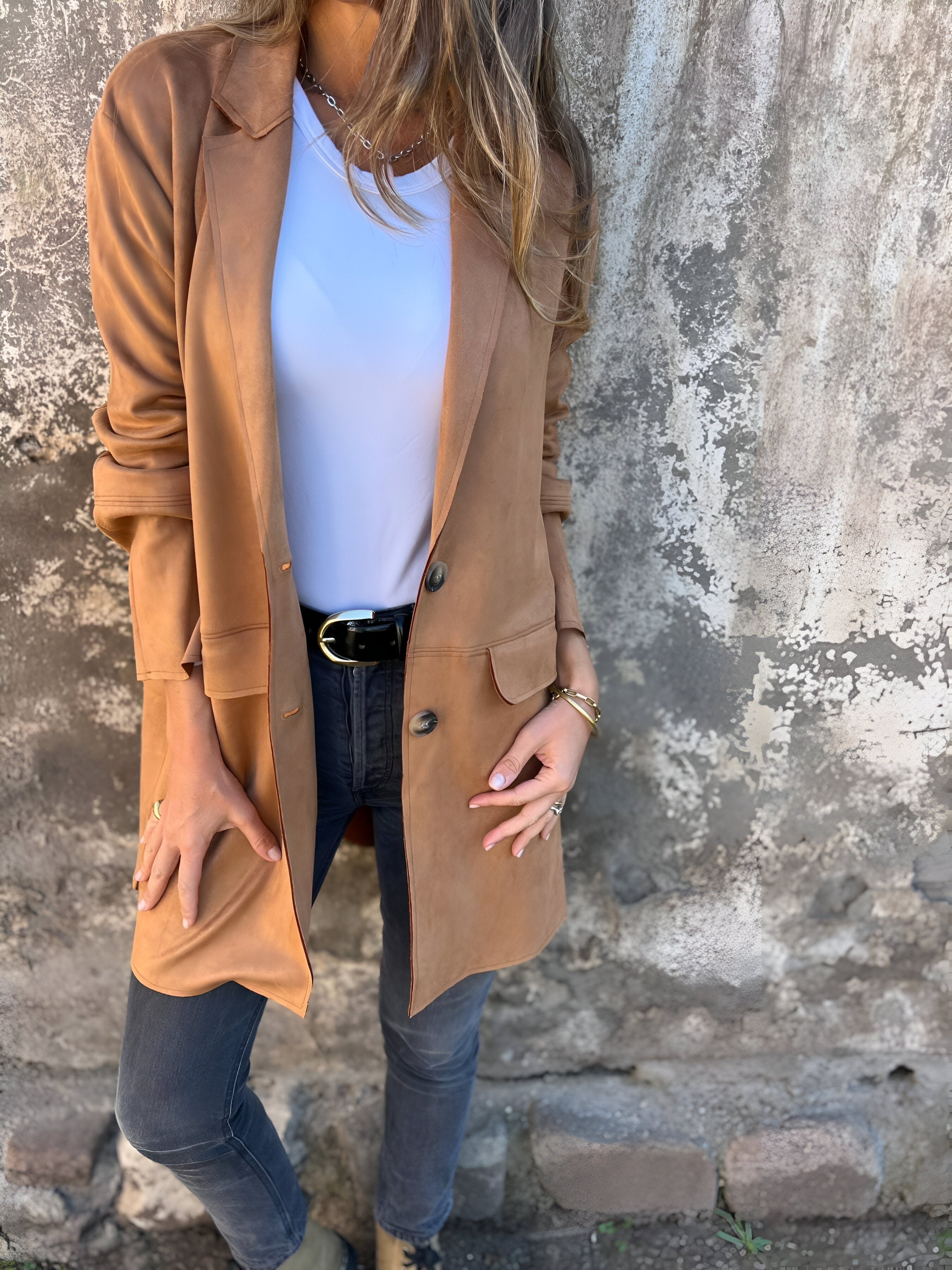 Donna con giacca lunga marrone, maglietta bianca e jeans neri, posa contro un muro rustico. Moda autunnale casual, stile urbano chic.