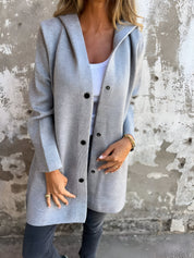 Donna indossa cappotto grigio con bottoni, su sfondo di muro rustico. Moda autunnale, abbigliamento casual elegante, stile urbano.