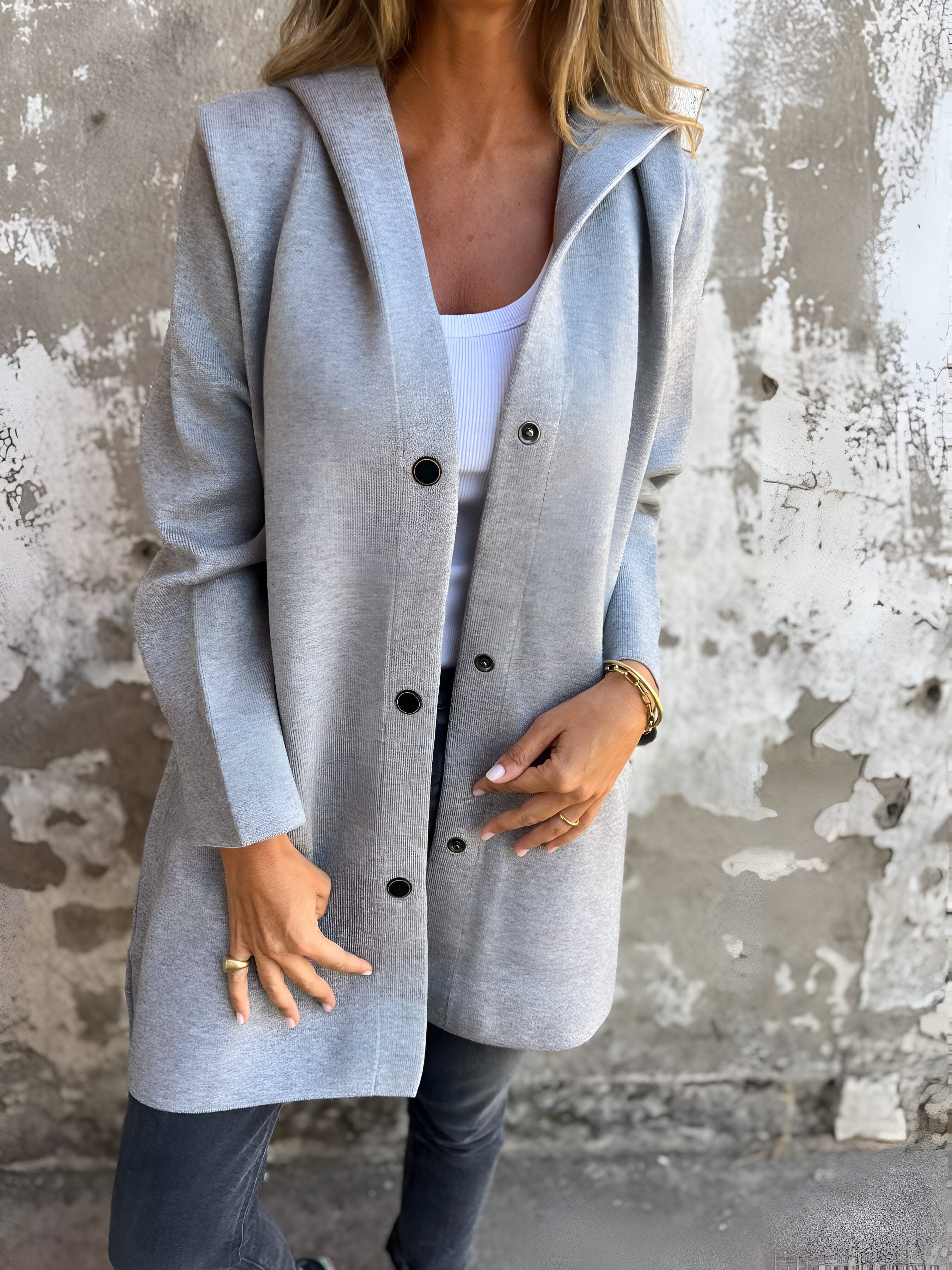 Donna indossa cappotto grigio con bottoni, su sfondo di muro rustico. Moda autunnale, abbigliamento casual elegante, stile urbano.
