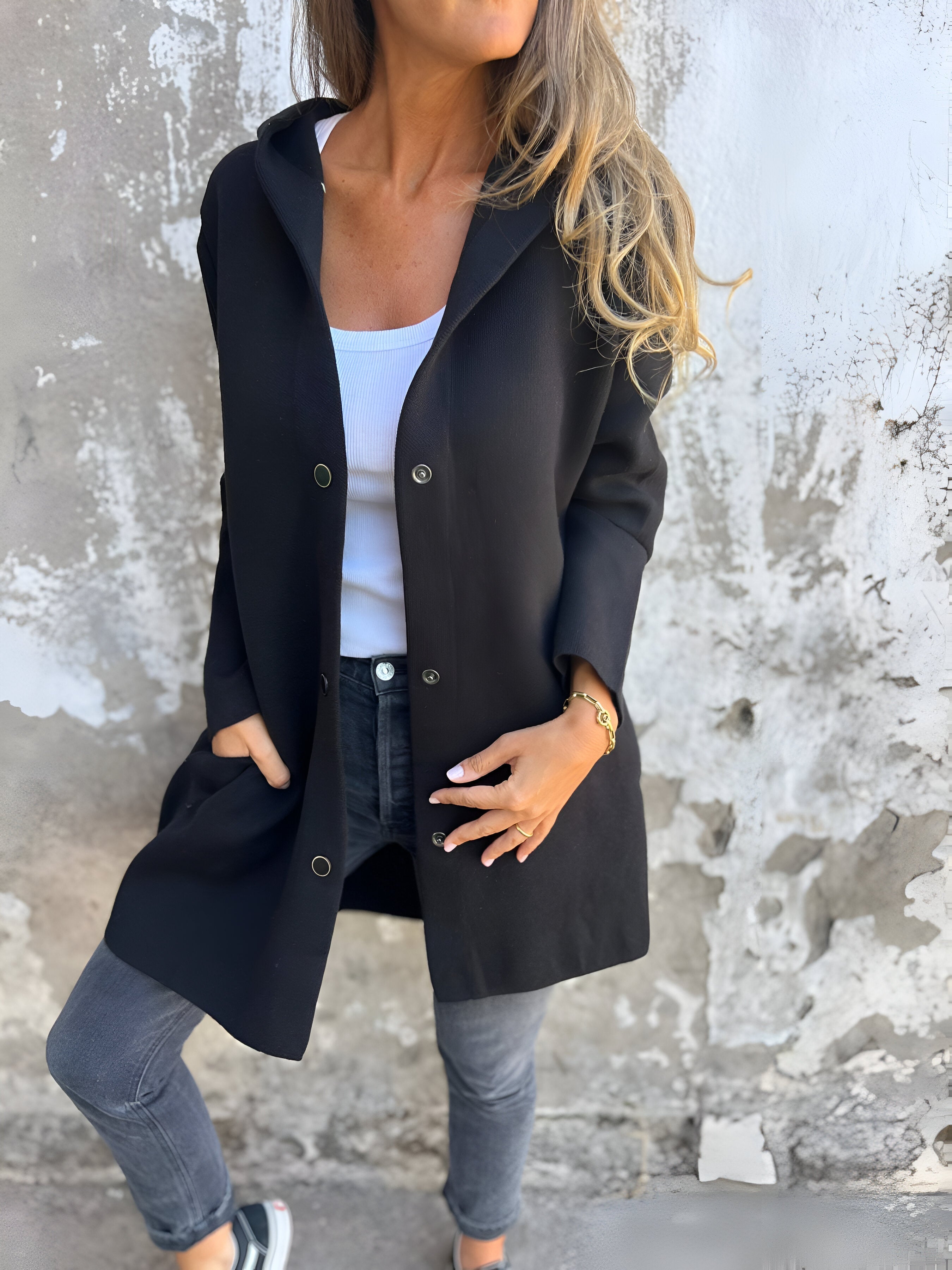Donna con cappotto nero elegante, jeans grigi e top bianco, posa contro muro rustico. Moda autunnale casual chic, stile urbano.