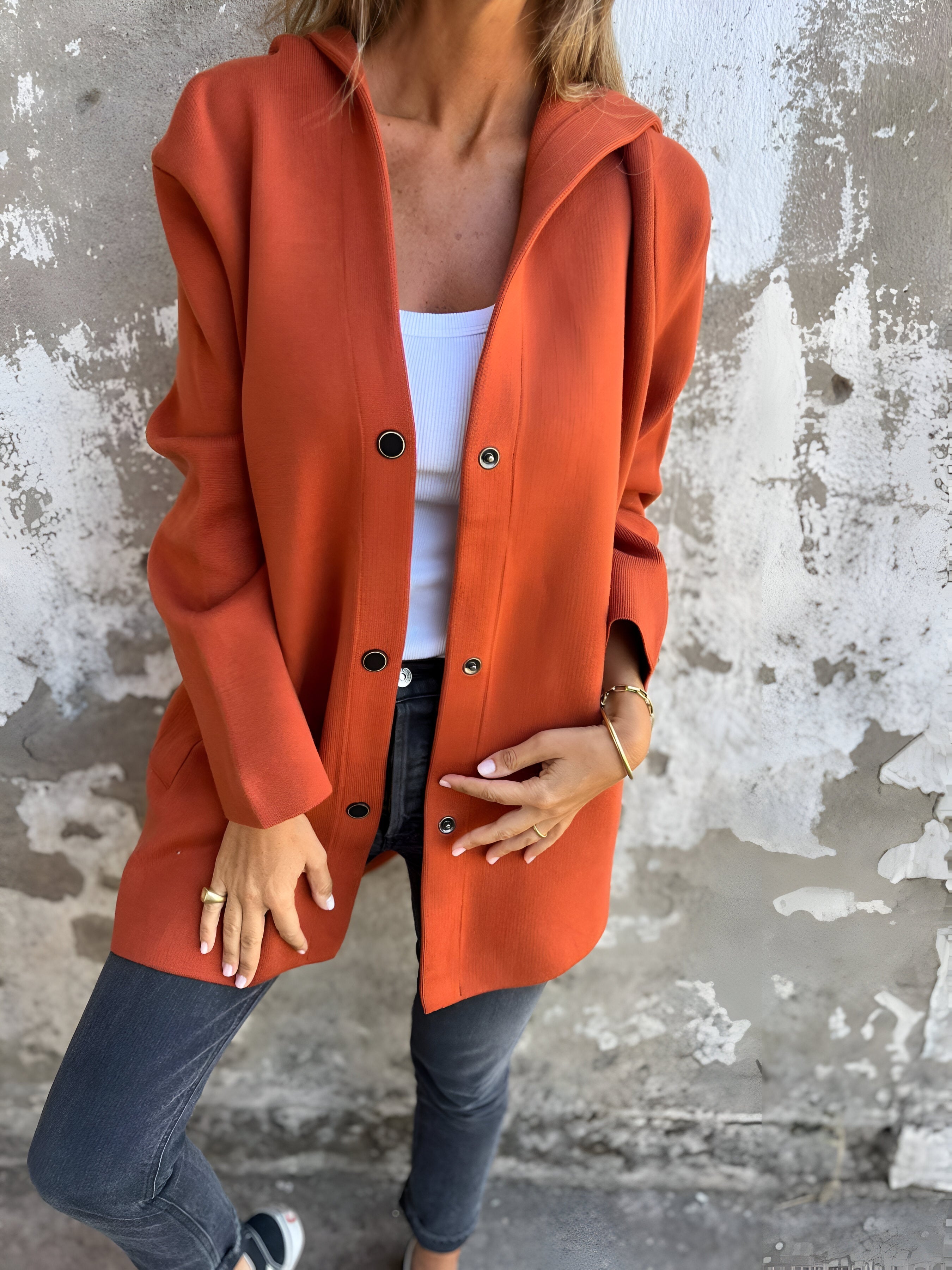 Giacca arancione donna, stile casual, abbinata a top bianco e jeans grigi. Moda autunnale, look urbano, sfondo muro rustico.