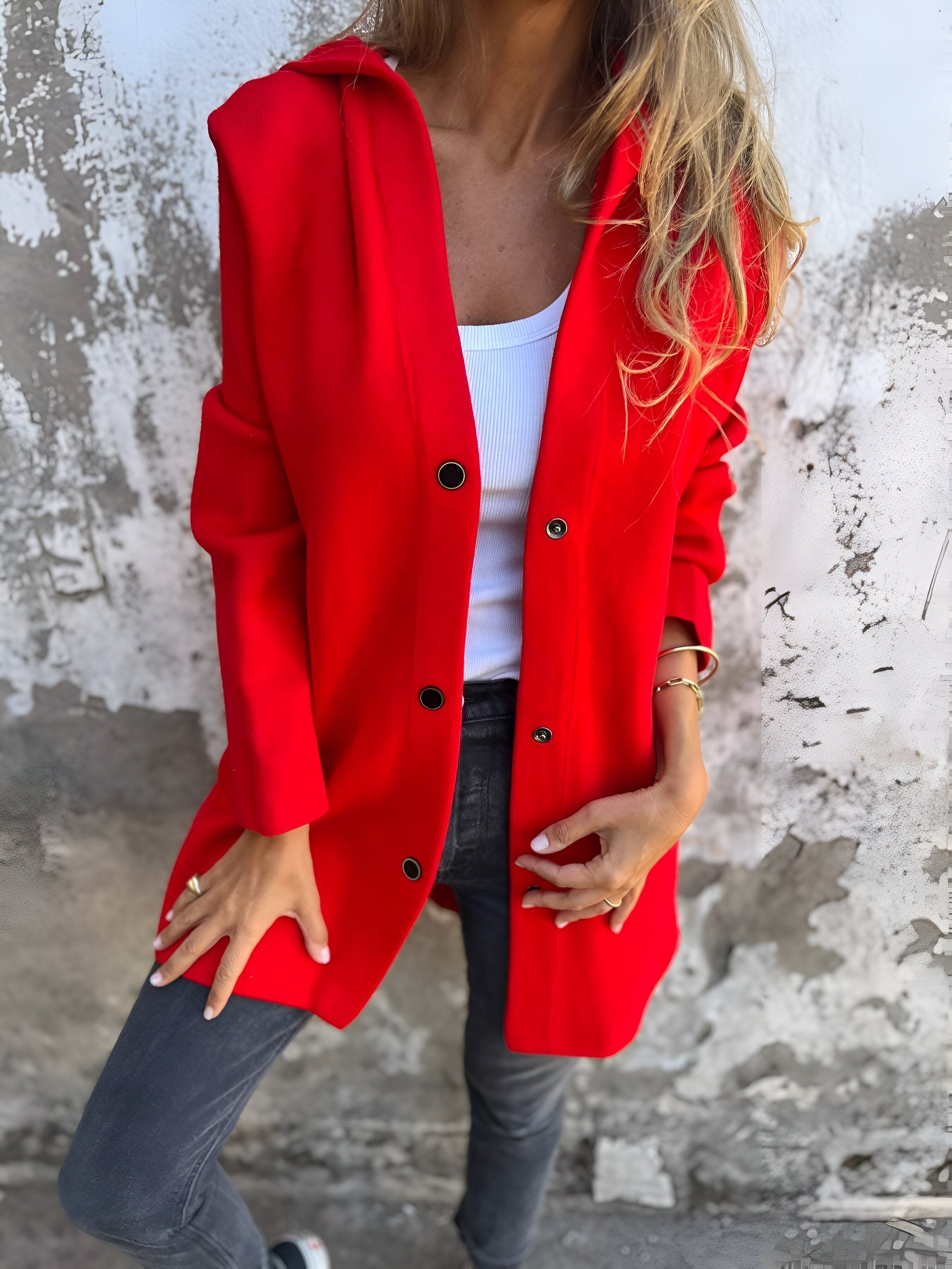Donna con cappotto rosso elegante, top bianco e jeans grigi, posa contro un muro rustico. Moda autunnale, stile casual chic, abbigliamento femminile.