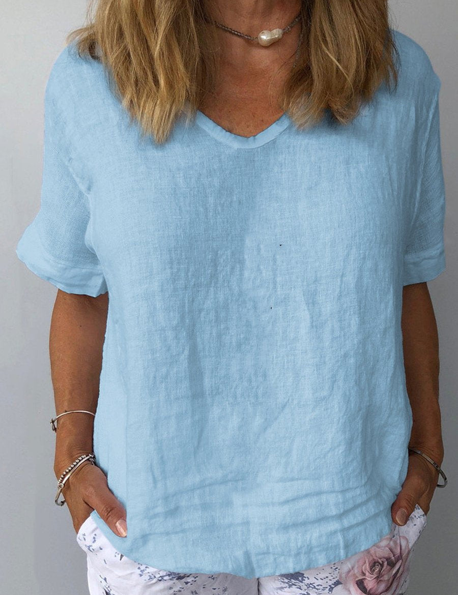 Donna con maglietta di lino azzurra, scollo a V, maniche corte, abbinata a pantaloni bianchi con stampa floreale. Moda estiva casual elegante.