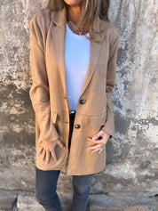 Donna con cappotto beige elegante, camicia bianca e jeans scuri, posa contro un muro rustico. Moda autunnale, stile casual chic.