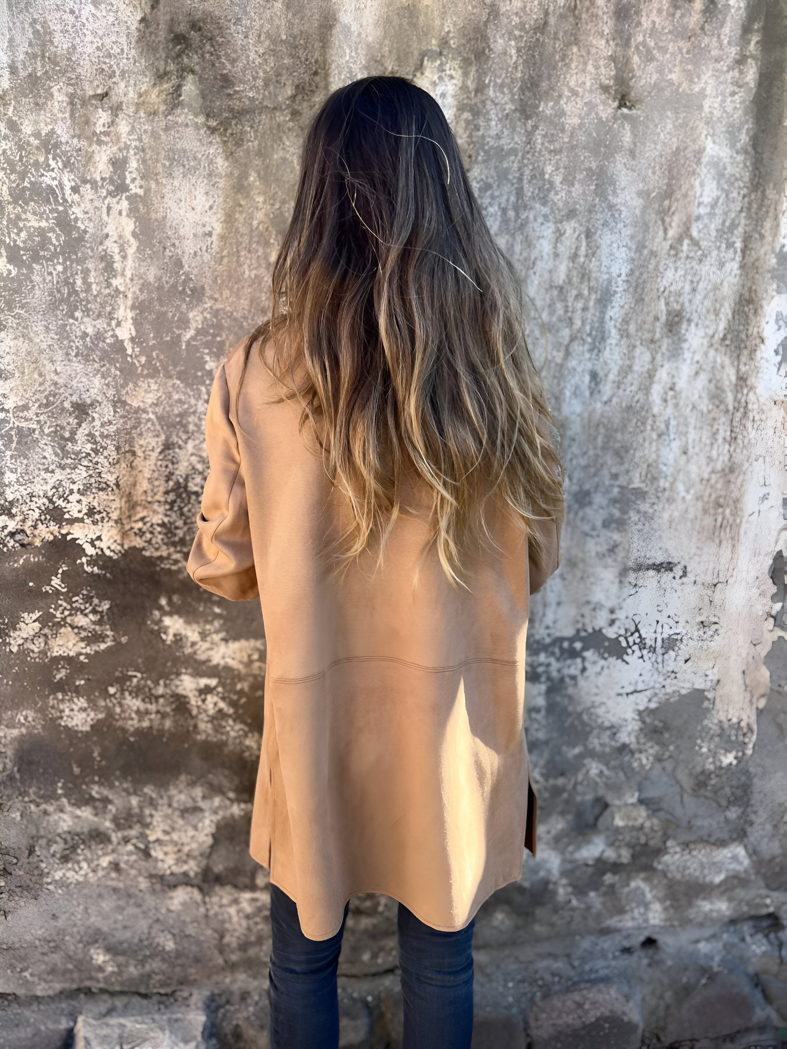Donna con cappotto beige e capelli lunghi davanti a un muro di cemento. Moda autunno, stile casual, abbigliamento femminile.