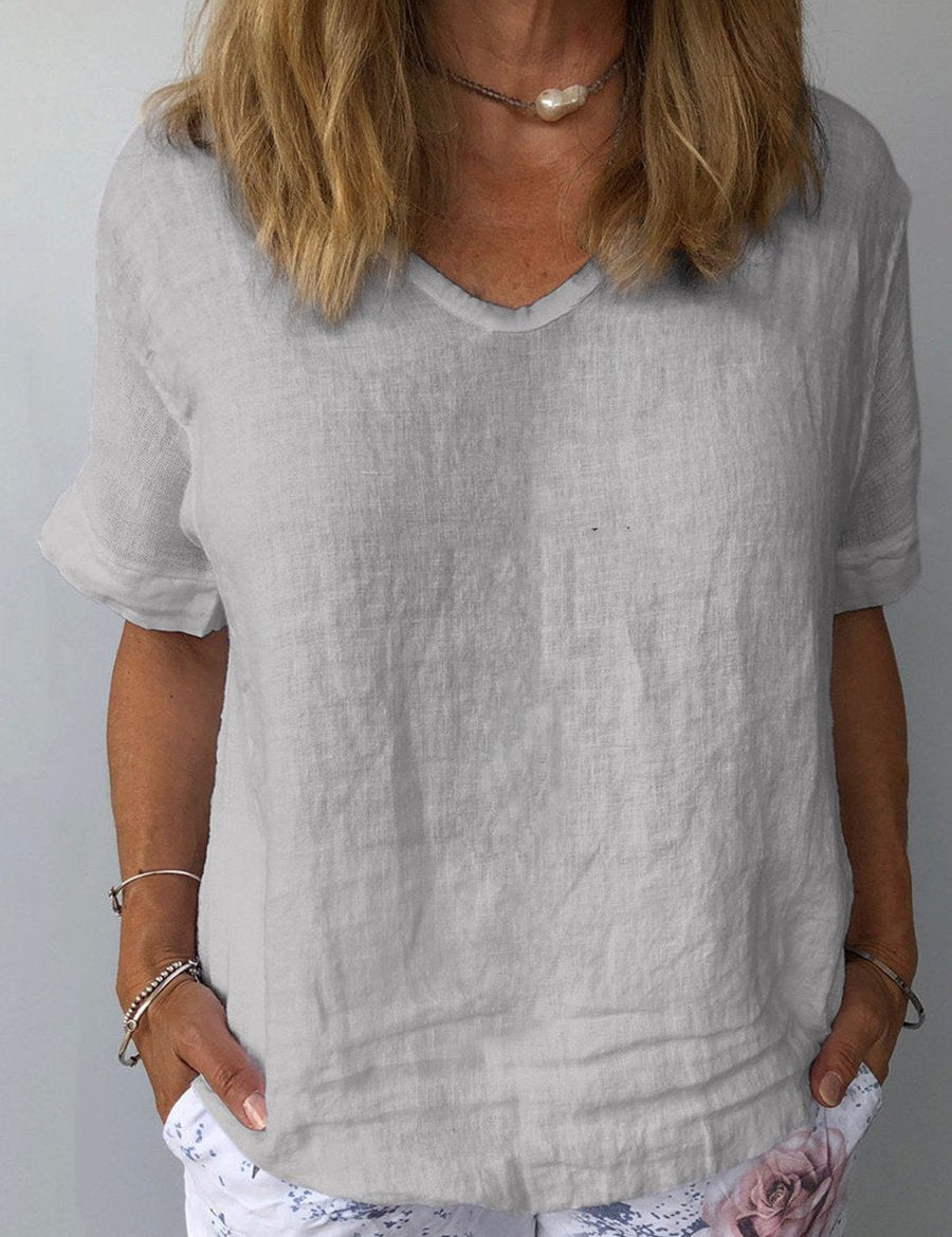 Donna con maglietta di lino grigia, scollo a V, maniche corte, indossa braccialetti e collana, pantaloni bianchi con stampa floreale.
