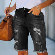 Shorts di jeans neri strappati da donna, lunghezza al ginocchio, con orlo sfrangiato. Abbinati a borsa marrone e maglietta bianca. Moda casual estiva.