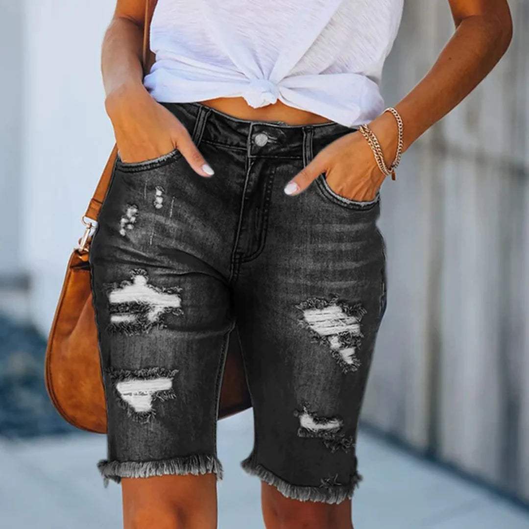 Shorts di jeans neri strappati da donna, lunghezza al ginocchio, con orlo sfrangiato. Abbinati a top bianco annodato e borsa marrone. Moda casual estiva.