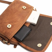 Borsa a tracolla in pelle marrone con chiusura a bottone, contenente un portafoglio nero e un power bank bianco. Design elegante e pratico.