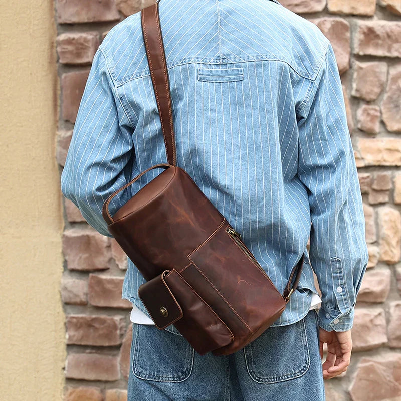 Uomo con giacca di jeans indossa borsa a tracolla in pelle marrone, design vintage, tasca frontale con bottone, sfondo muro di mattoni.