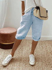 Donna con pantaloncini di lino azzurri, maglietta bianca, borsa a tracolla beige e sneakers bianche su tappeto intrecciato. Moda casual estiva.