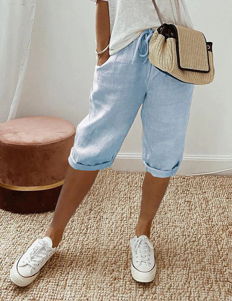 Donna con pantaloncini di lino azzurri, maglietta bianca, borsa a tracolla beige e sneakers bianche su tappeto intrecciato. Moda casual estiva.