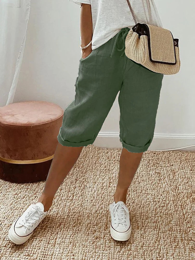 Donna con pantaloncini verdi, maglietta bianca, borsa a tracolla beige e sneakers bianche su tappeto intrecciato. Moda casual estiva.
