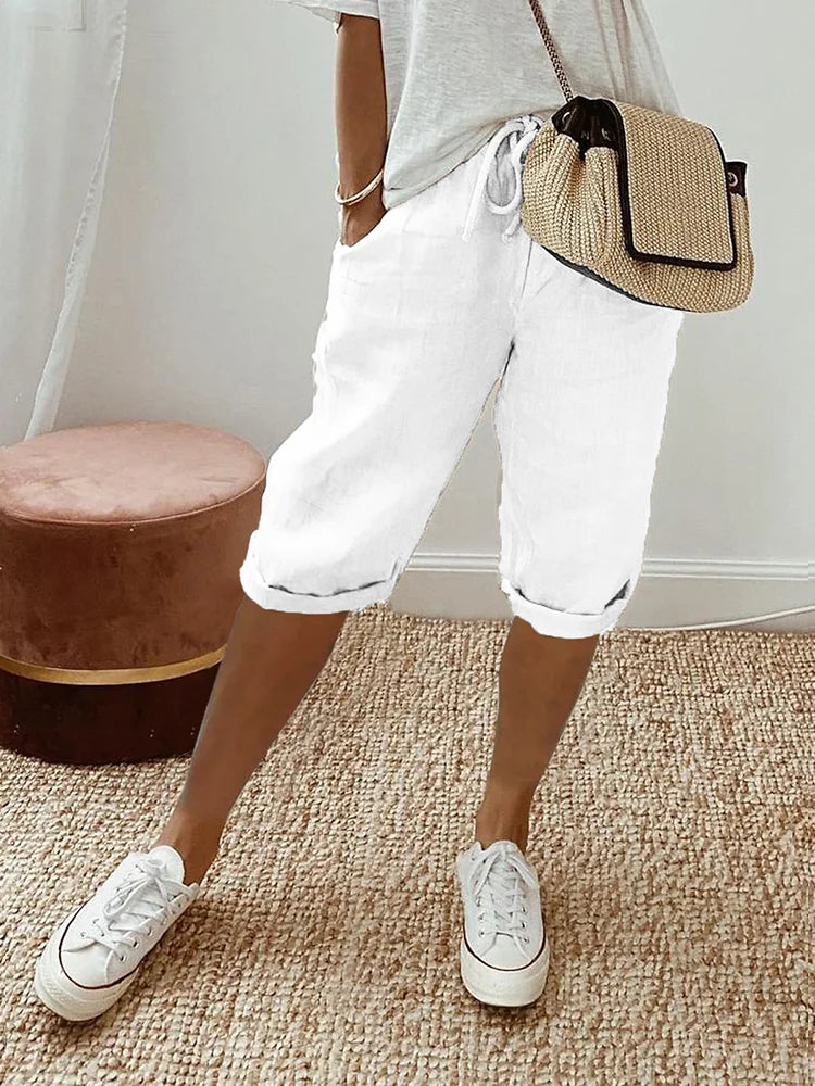 Donna con pantaloncini bianchi casual, maglietta grigia, borsa a tracolla beige e sneakers bianche su tappeto beige. Moda estiva, abbigliamento casual.
