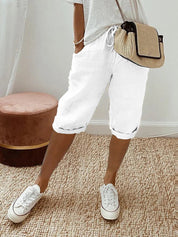 Donna con pantaloncini bianchi casual, maglietta grigia, borsa a tracolla beige e sneakers bianche su tappeto beige. Moda estiva, abbigliamento casual.