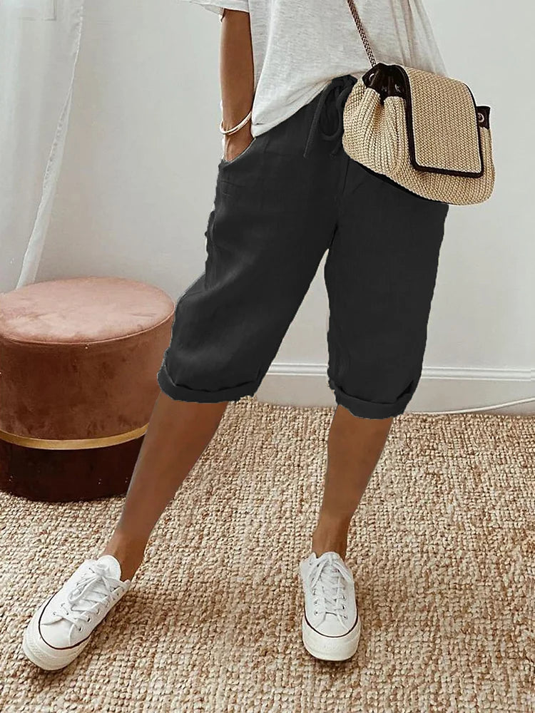 Pantaloncini neri casual da donna, abbinati a maglietta bianca e borsa a tracolla beige, su tappeto intrecciato. Scarpe da ginnastica bianche.