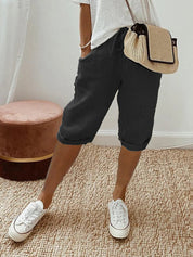 Pantaloncini neri casual da donna, abbinati a maglietta bianca e borsa a tracolla beige, su tappeto intrecciato. Scarpe da ginnastica bianche.