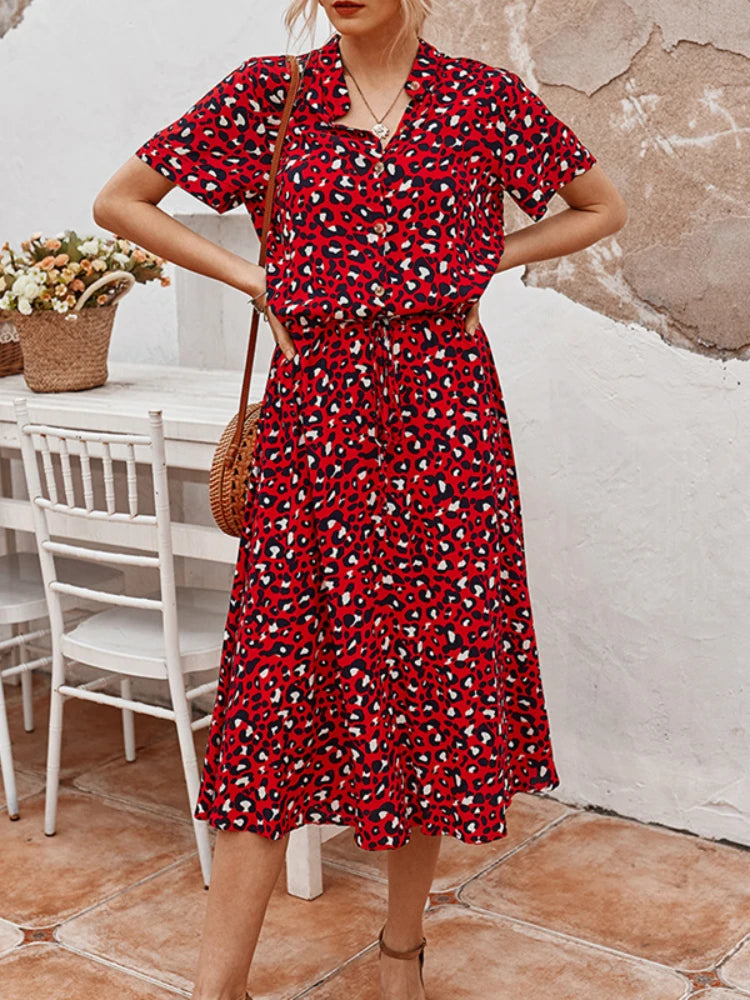 Abito rosso a fantasia leopardata, maniche corte, lunghezza media, indossato da donna. Sfondo con tavolo bianco e cesto di fiori.
