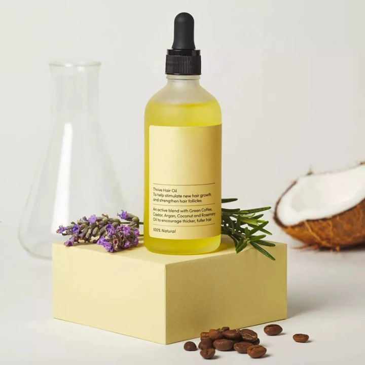 Bottiglia di olio per capelli su piedistallo, circondata da lavanda, rosmarino, chicchi di caffè e cocco. Cura capelli naturale, crescita e rinforzo.