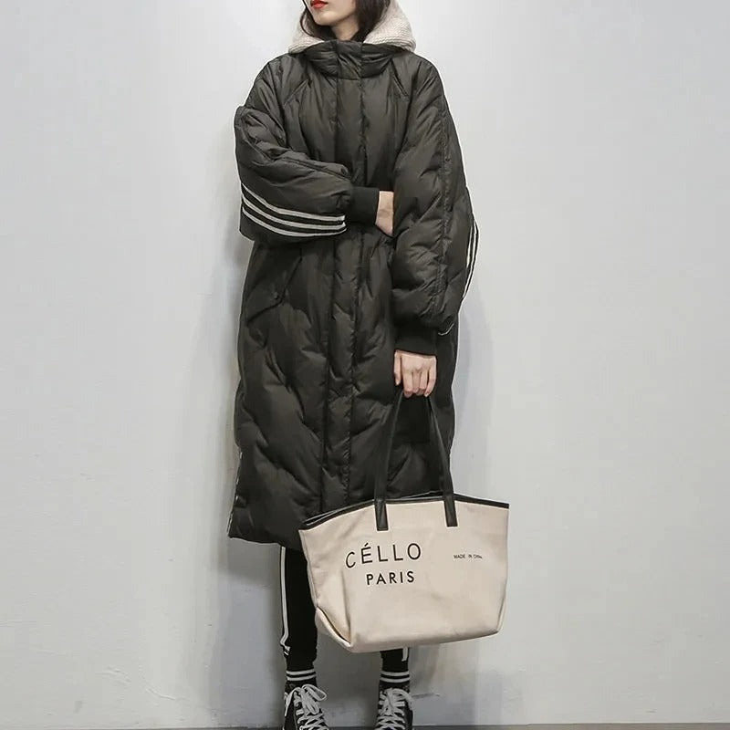 Cappotto lungo nero imbottito con cappuccio, dettagli a righe bianche sulle maniche, abbinato a borsa beige "Céllo Paris" e pantaloni neri.