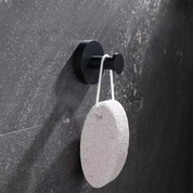 Pietra pomice bianca appesa a un gancio nero su parete grigia, ideale per esfoliazione piedi e cura della pelle. Accessorio bagno essenziale.