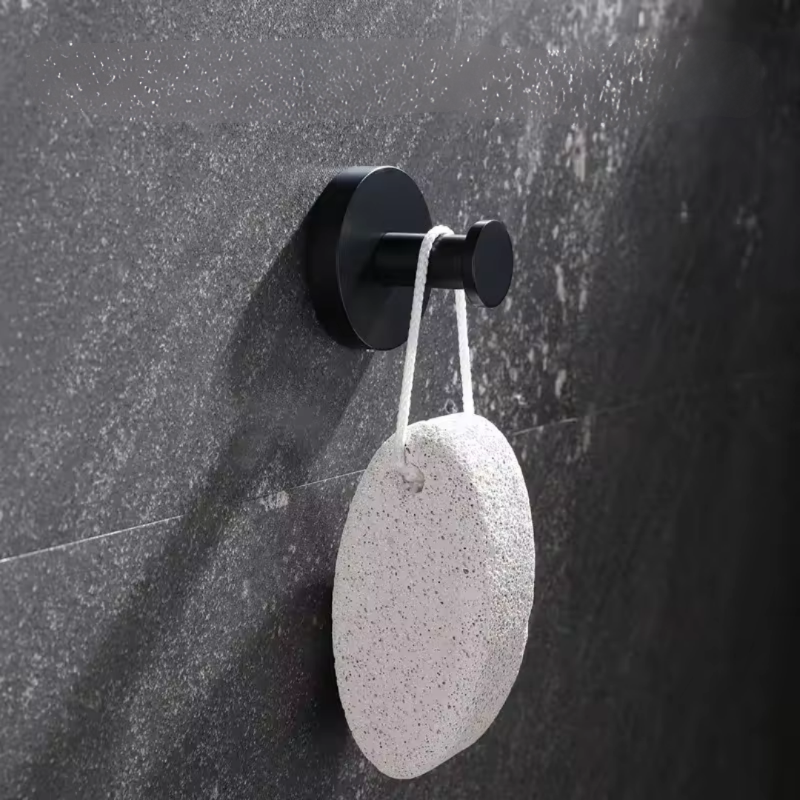 Pietra pomice bianca appesa a un gancio nero su parete grigia, ideale per esfoliazione piedi e cura della pelle. Accessorio bagno essenziale.