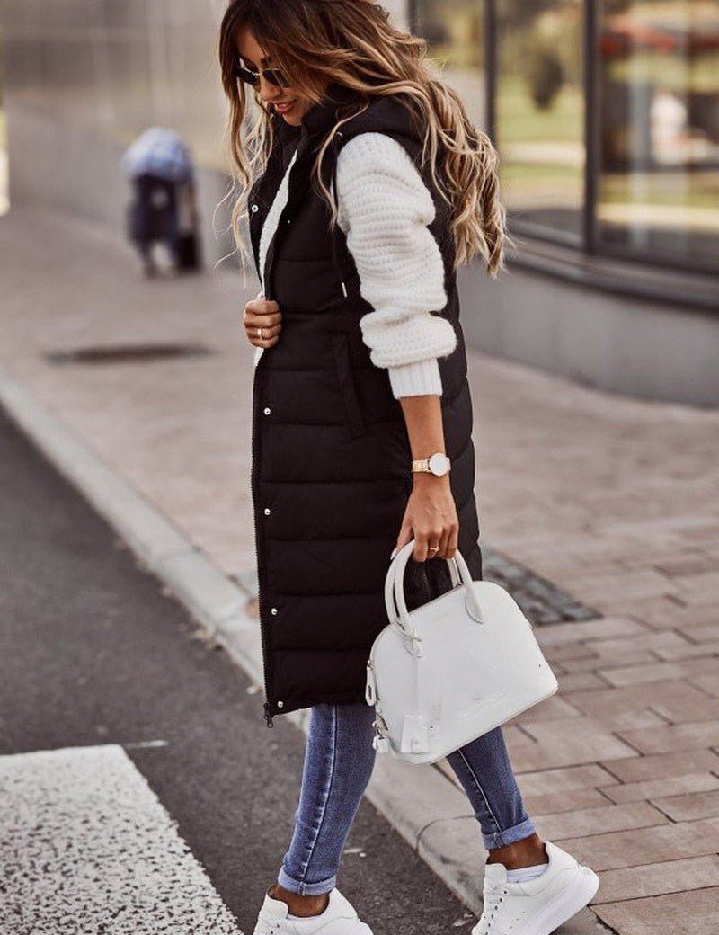 Donna con gilet lungo nero imbottito, maglione bianco, jeans aderenti e sneakers bianche, borsa bianca. Moda autunnale casual chic.