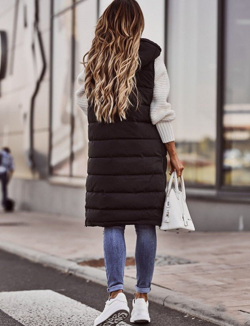 Donna di spalle con gilet lungo nero imbottito, jeans aderenti, sneakers bianche e borsa bianca. Moda autunnale casual in città.