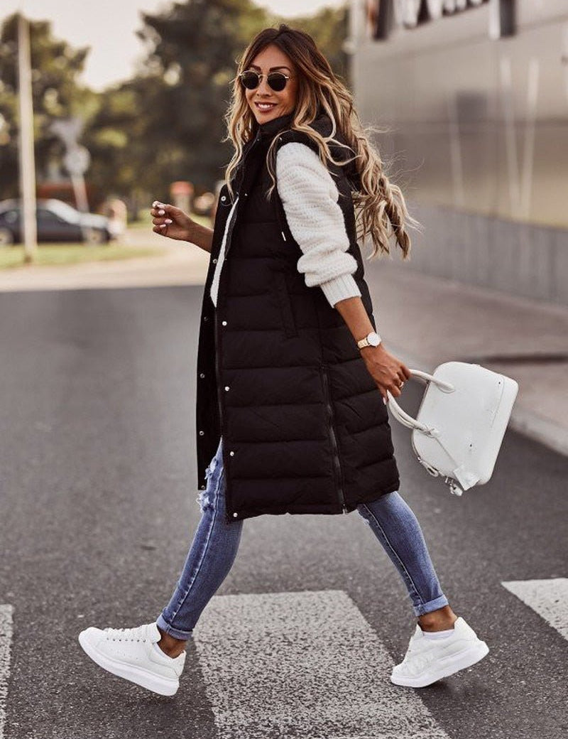 Donna alla moda con gilet lungo nero, jeans attillati e sneakers bianche, attraversa la strada con borsa bianca. Look casual autunnale.