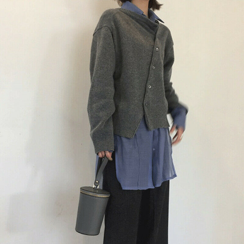 Maglione grigio asimmetrico su camicia blu, pantaloni neri, borsa a cilindro grigia. Moda casual autunnale, abbigliamento donna elegante.