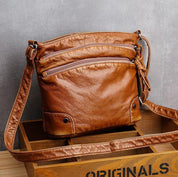 Borsa a tracolla in pelle marrone, design vintage, con tasche frontali con zip, su sfondo grigio. Accessorio moda donna elegante e pratico.