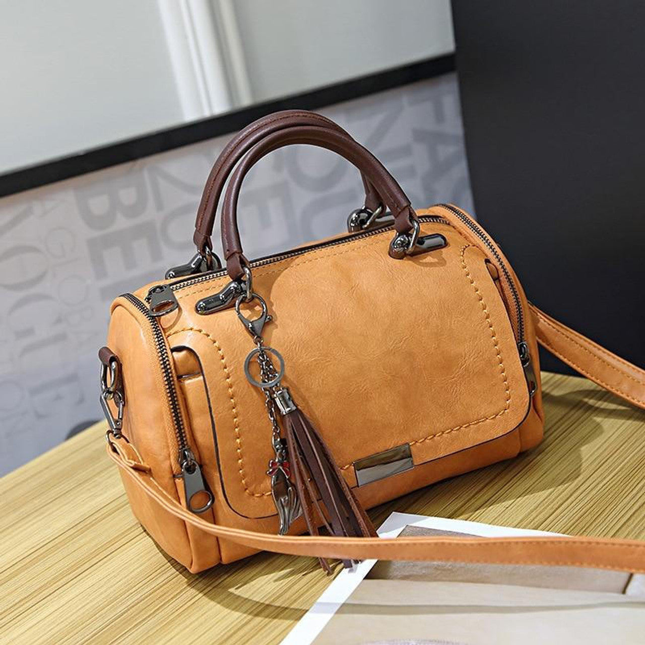 Borsa a tracolla in pelle marrone chiaro con manici, dettagli in metallo e ciondolo con frange, su tavolo di legno. Elegante e versatile.