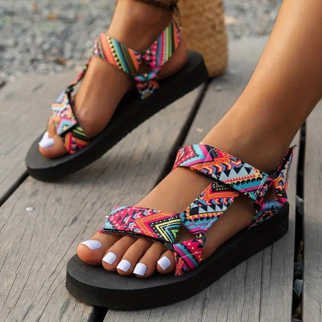 Sandali estivi donna con suola nera e cinturini multicolore a motivi geometrici, indossati su piedi con smalto bianco, su pavimento in legno.