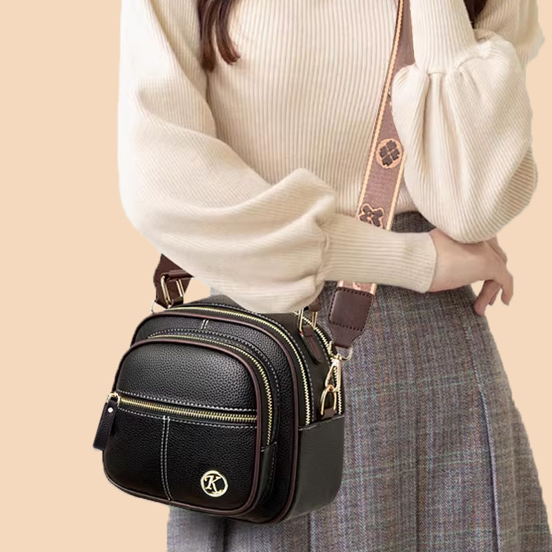 Borsa a tracolla in pelle nera con dettagli dorati, indossata da una donna in maglione beige e gonna grigia. Accessorio moda elegante e versatile.