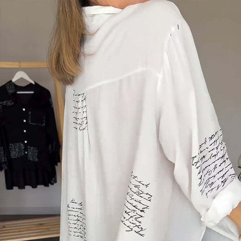 Camicia bianca oversize con scritte nere, maniche lunghe, tessuto leggero. Sullo sfondo, camicia nera simile appesa. Moda casual elegante.