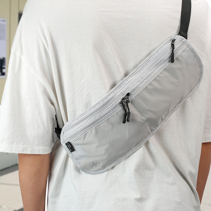 Zaino monospalla grigio chiaro indossato su maglietta bianca, con doppia cerniera e design minimalista. Accessorio moda unisex, pratico e versatile.