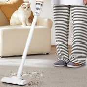 Aspirapolvere senza fili bianco su tappeto beige, accanto a persona in pigiama a righe e gatto bianco su divano. Pulizia casa, elettrodomestico moderno.