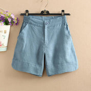 Pantaloncini donna in lino blu, elastico in vita, tasche laterali, appesi su gruccia nera. Design casual estivo, sfondo beige, fiori decorativi.