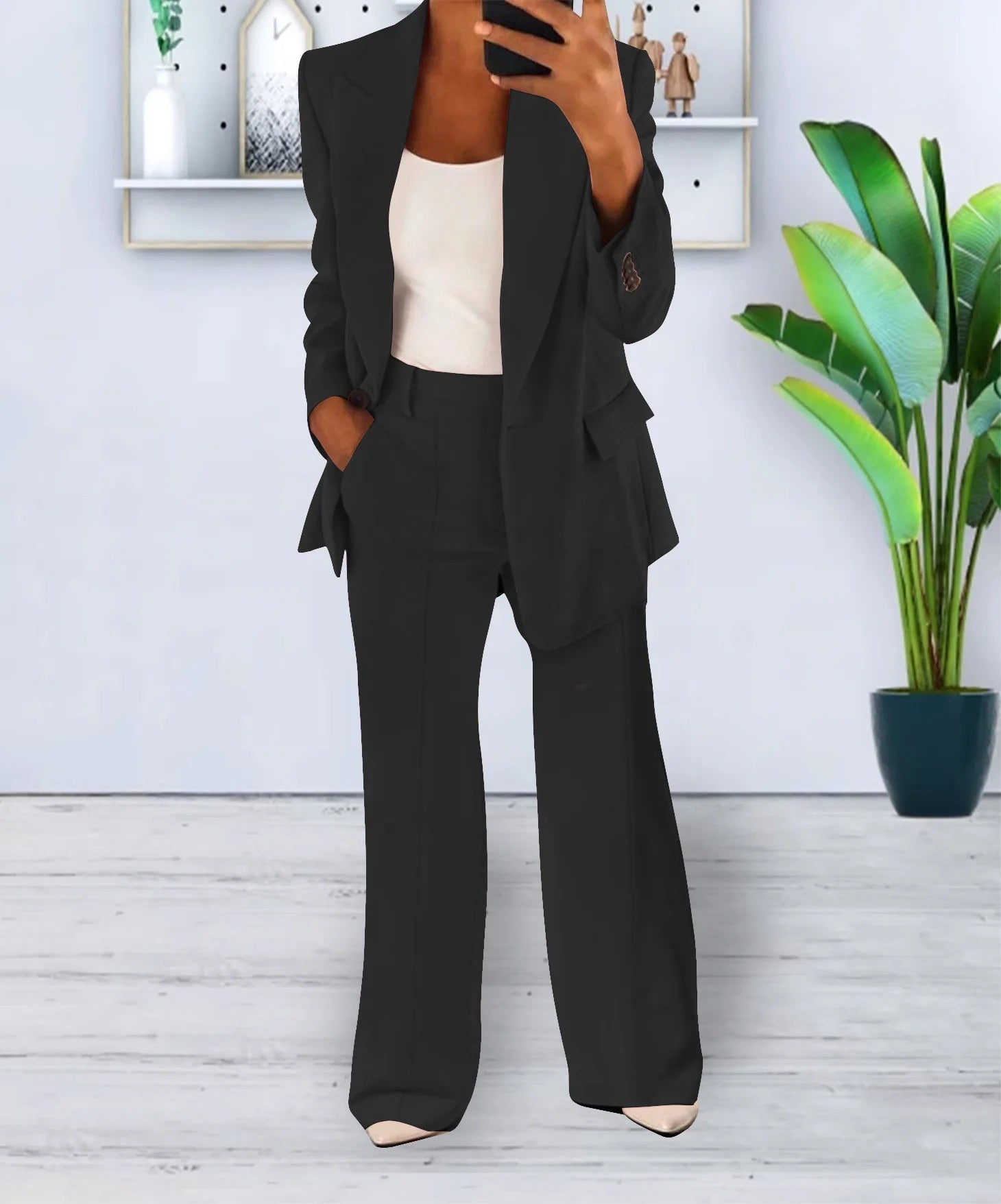 Donna in elegante completo nero, giacca e pantaloni, con top bianco. Sfondo moderno con pianta verde. Moda femminile, abbigliamento formale.