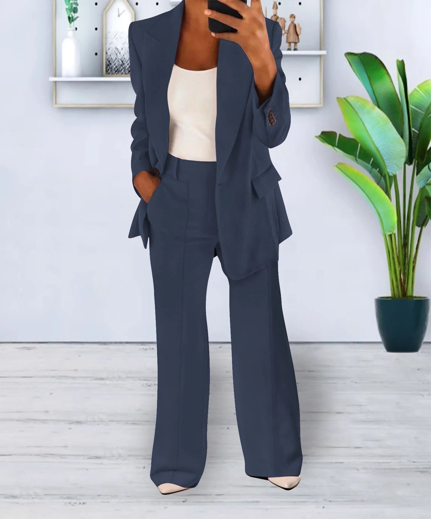Completo donna elegante blu scuro, giacca e pantaloni a zampa, indossato con top bianco. Sfondo moderno con pianta decorativa.