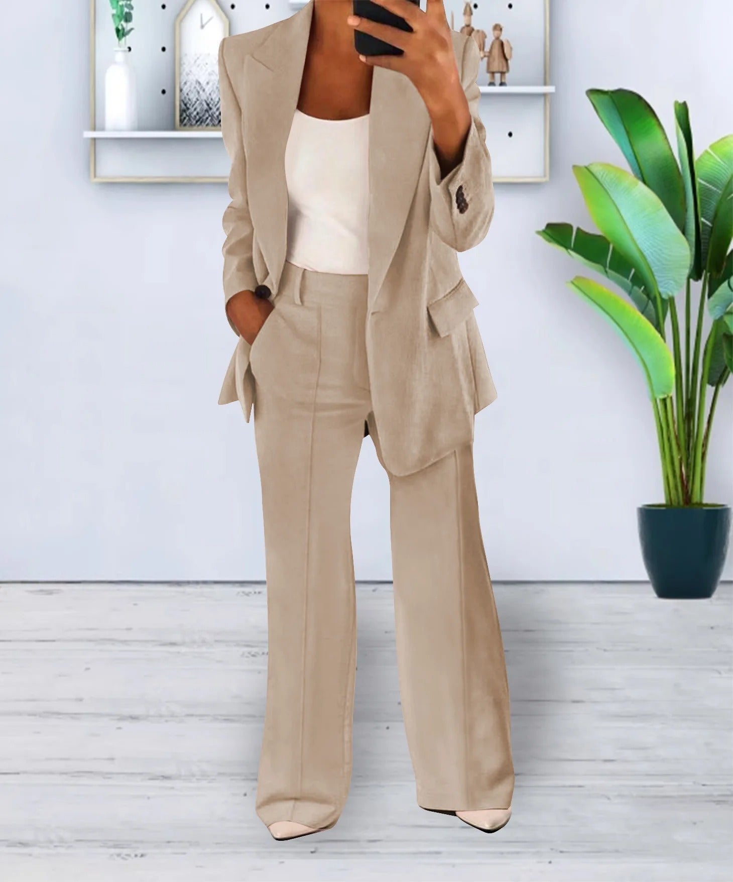 Completo donna beige elegante, giacca e pantaloni a zampa, indossato con top bianco. Sfondo moderno con pianta decorativa. Moda femminile chic.