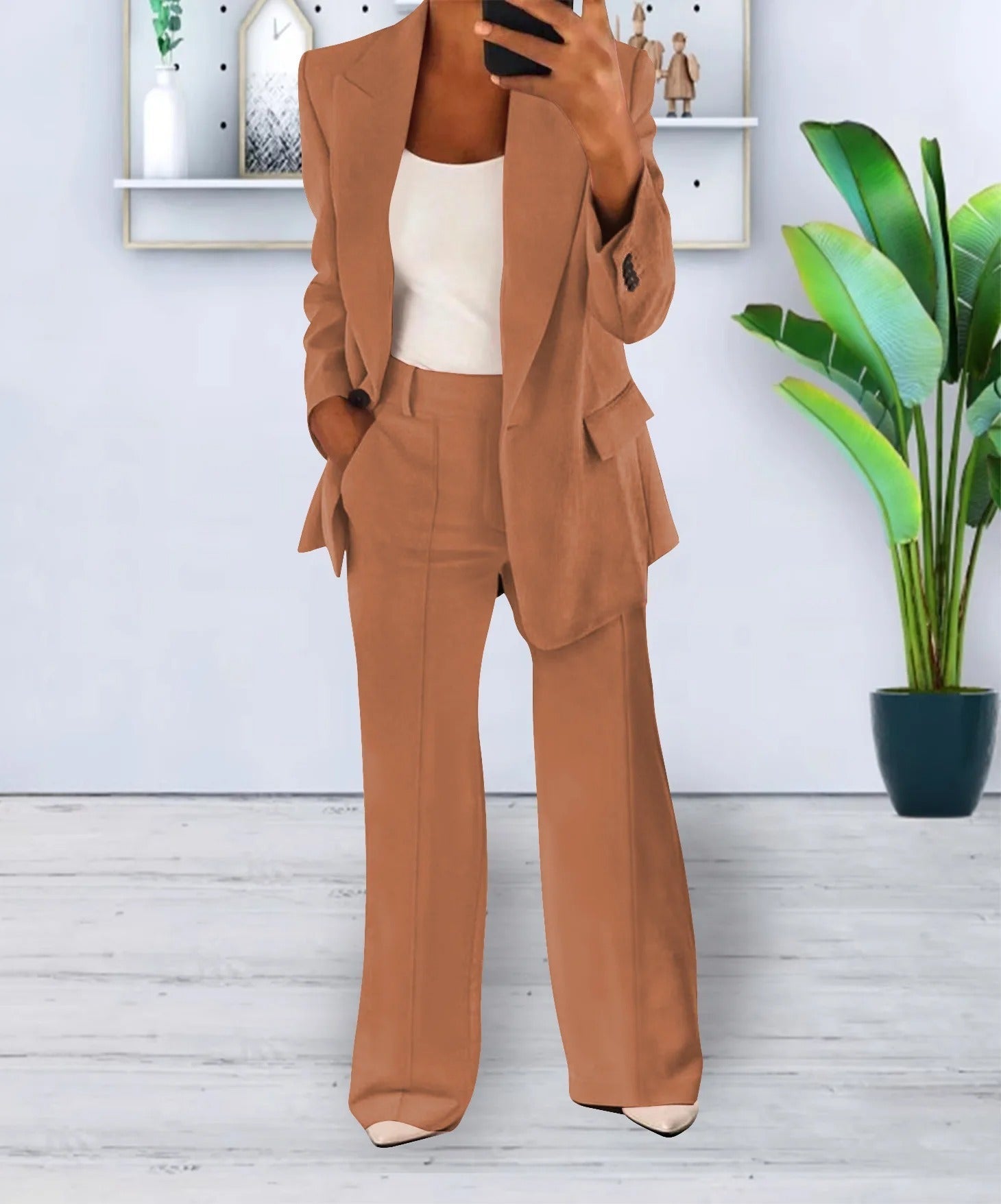 Completo donna elegante marrone, giacca e pantaloni a zampa, indossato con top bianco. Sfondo con pianta verde e mensola decorativa.