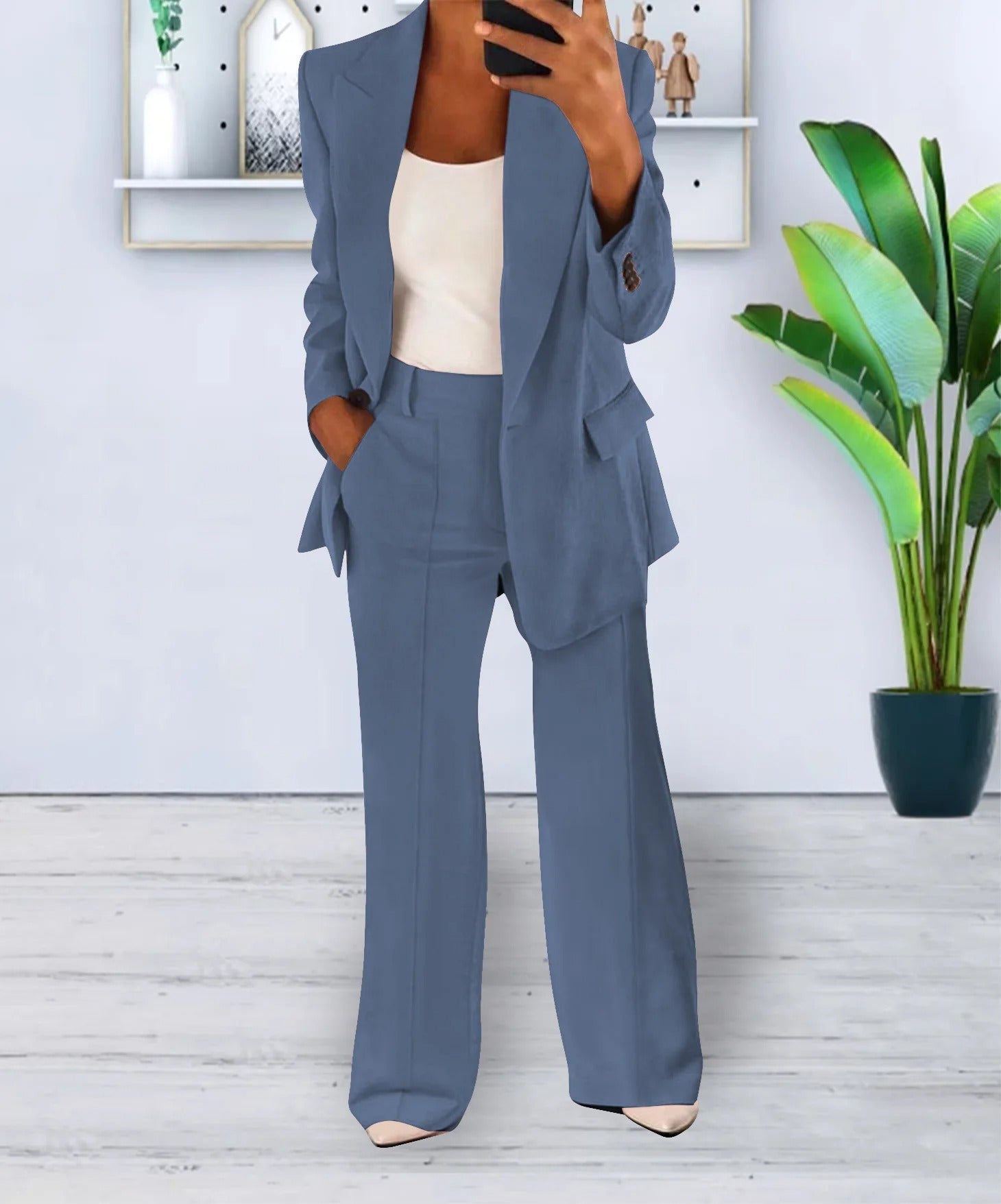 Completo donna elegante blu, giacca e pantaloni a zampa, indossato con top bianco. Sfondo moderno con pianta verde e mensola decorativa.