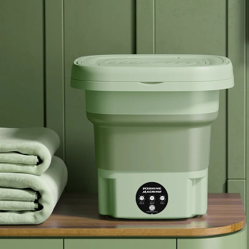 Lavatrice portatile pieghevole verde su tavolo di legno, accanto a asciugamani. Design compatto, ideale per piccoli spazi e viaggi.