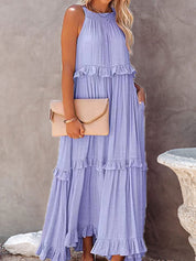 Abito lungo estivo viola con balze, senza maniche, indossato da donna bionda. Accessoriato con pochette beige. Moda femminile elegante.