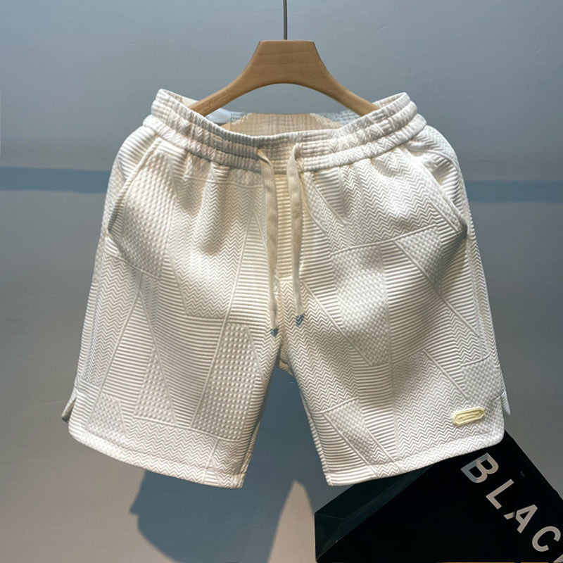 Pantaloncini bianchi in tessuto strutturato, appesi su gruccia di legno. Design geometrico, elastico in vita, ideale per moda estiva casual.