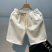 Pantaloncini bianchi in tessuto strutturato, appesi su gruccia di legno. Design geometrico, elastico in vita, ideale per moda estiva casual.